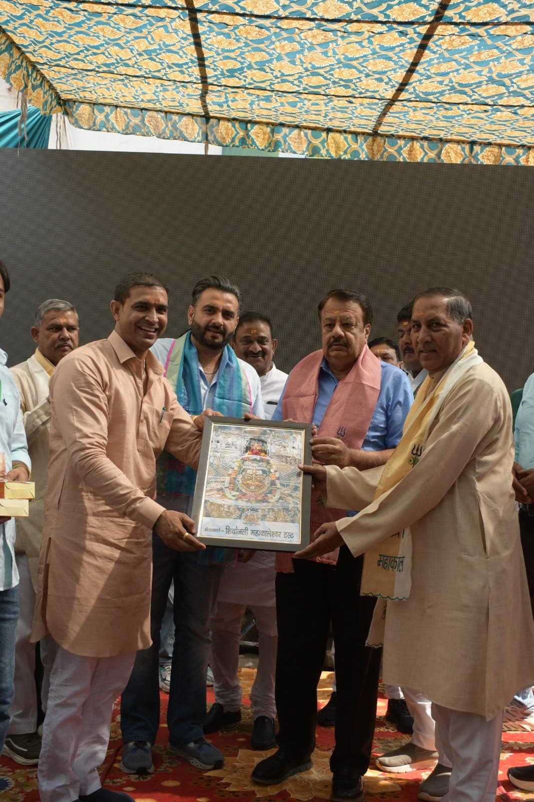 मंच पर चार पुरुष मिलकर महाकालेश्वर का फ्रेम किया हुआ चित्र पकड़े हैं, कई अंगवस्त्र पहने हैं