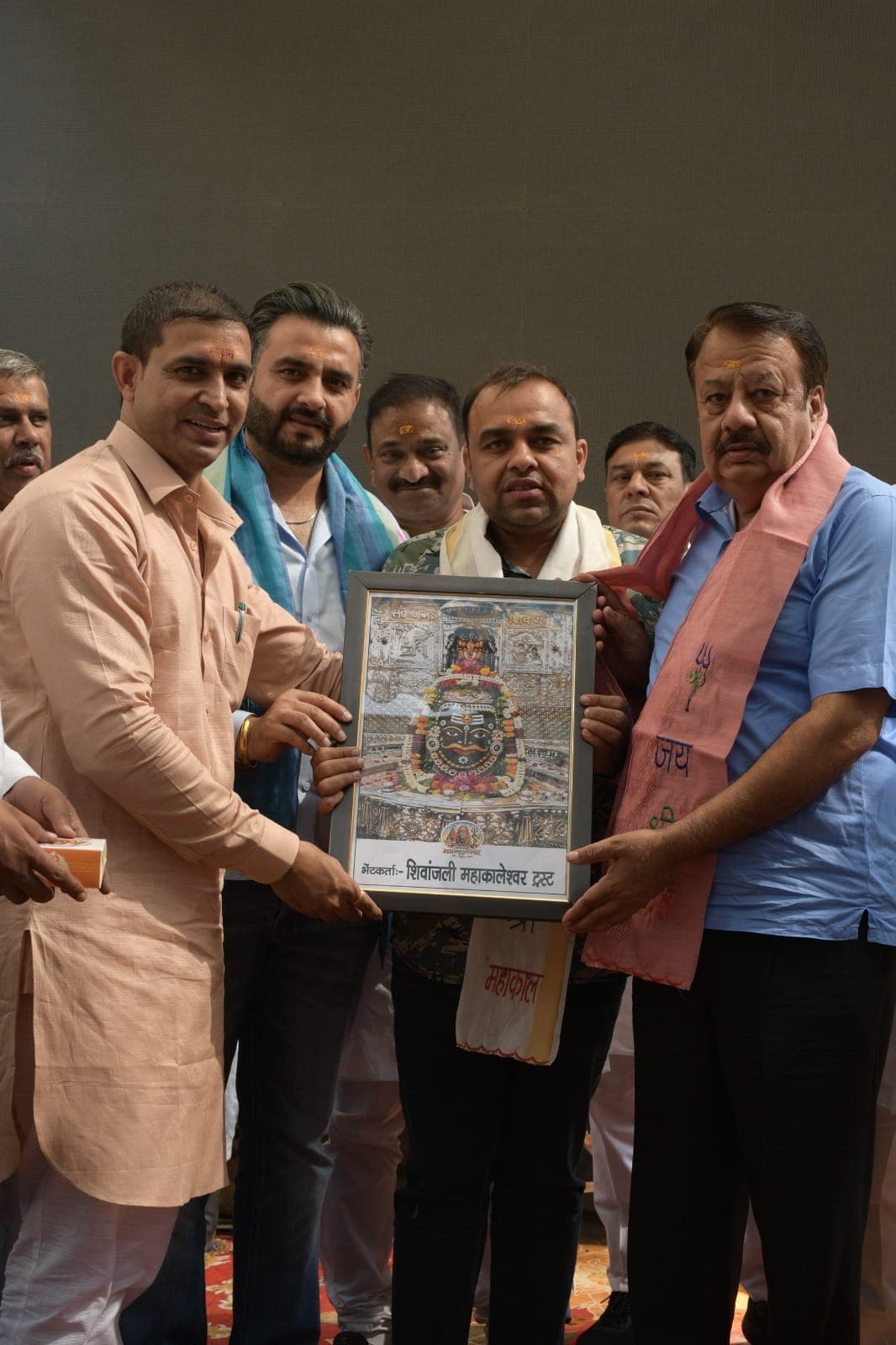 मंच पर अंगवस्त्र पहने अतिथि और कुर्ता पहने एक सदस्य महाकालेश्वर का फ्रेम किया हुआ चित्र थामे हैं