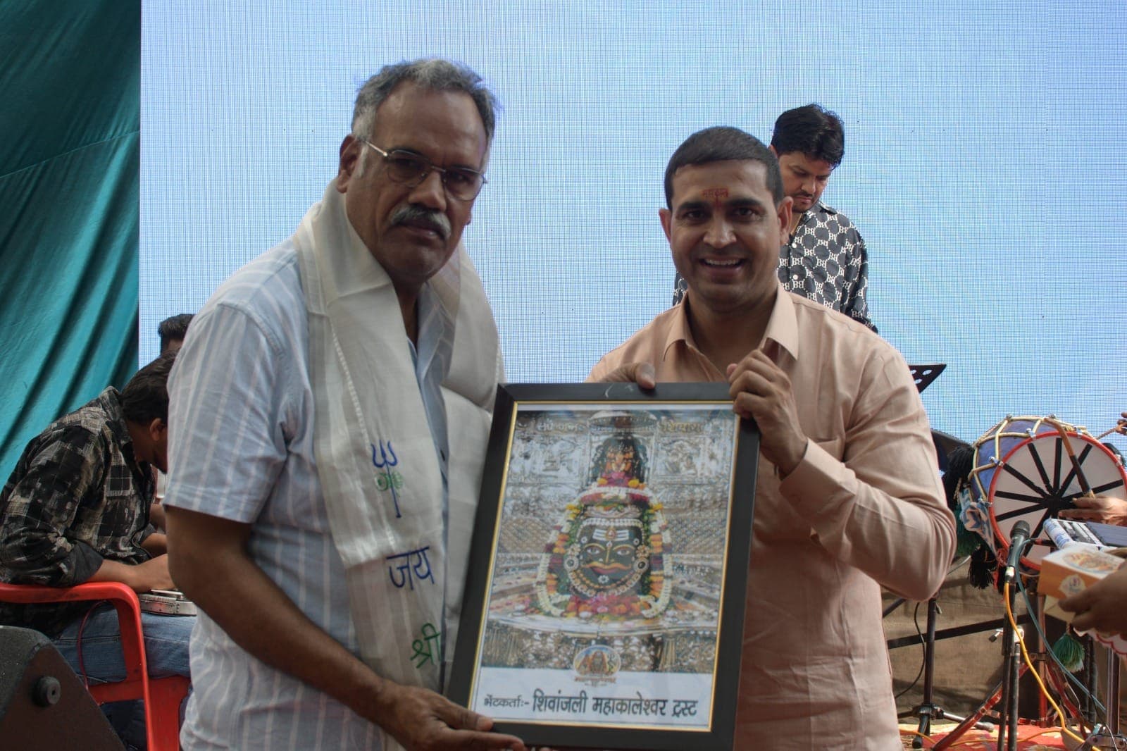 चश्मा पहने एक अतिथि, हल्के नारंगी कुर्ते वाले व्यक्ति के साथ महाकाल की फ्रेम की हुई तस्वीर थामे हुए।