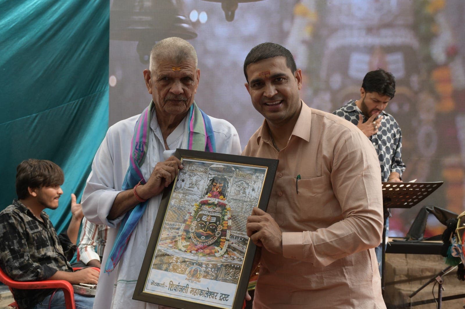 नीले-गुलाबी दुपट्टे वाले बुज़ुर्ग अतिथि, हल्के नारंगी कुर्ते वाले व्यक्ति के साथ महाकाल की तस्वीर थामे हुए।
