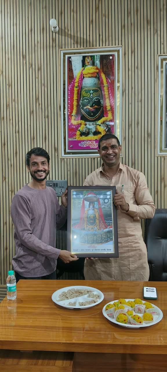 चश्मे और हल्के नीले दुपट्टे में एक अतिथि, हल्के नारंगी कुर्ते वाले व्यक्ति के साथ महाकाल की तस्वीर थामे।