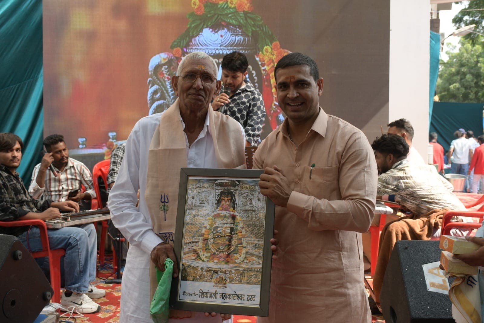 शॉल ओढ़े बुज़ुर्ग को हल्के नारंगी कुर्ते वाले सज्जन शिव जी की फ्रेम की तस्वीर भेंट करते हुए