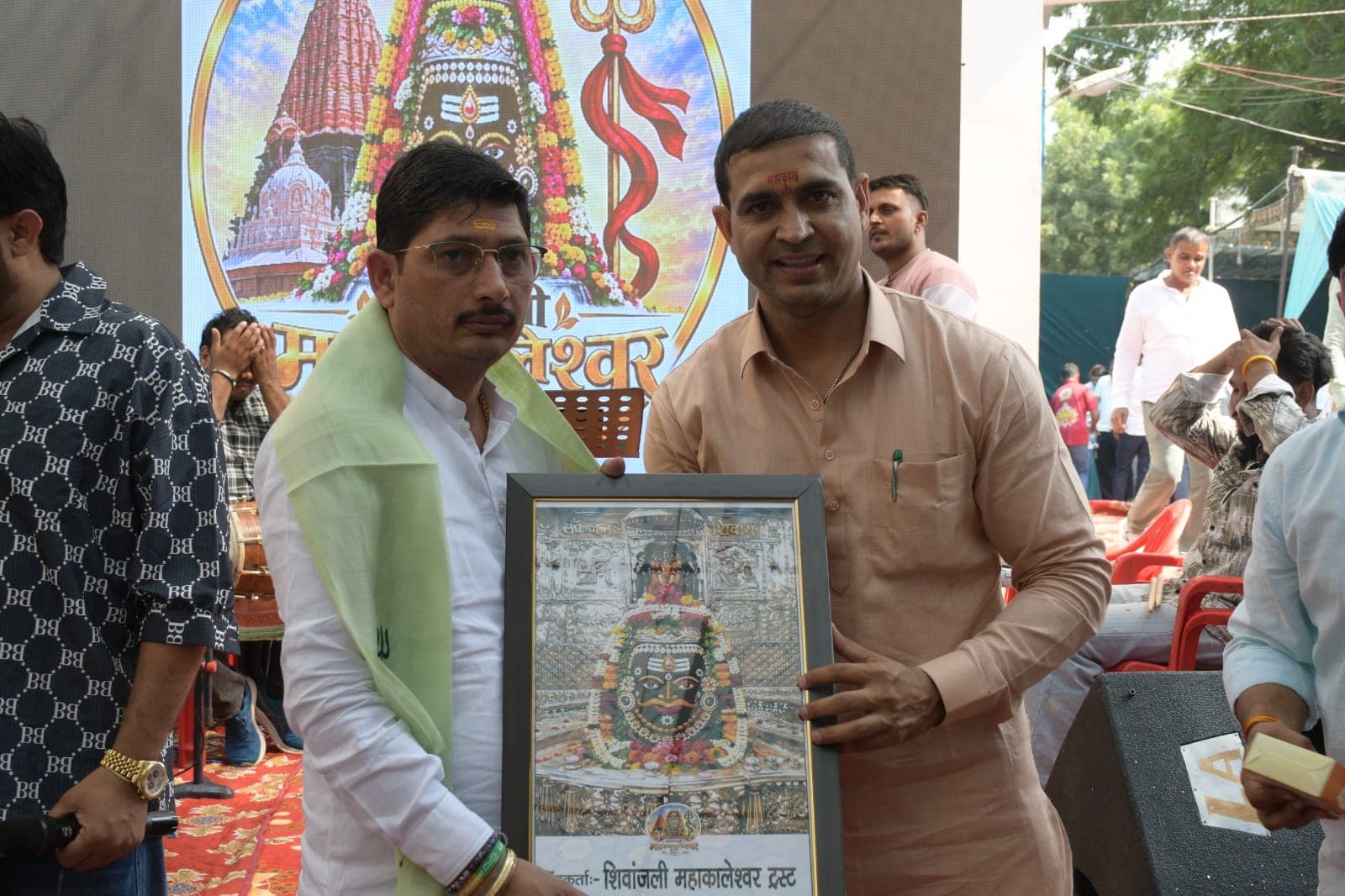 हरी शॉल पहने अतिथि को हल्के नारंगी कुर्ते वाले सज्जन शिव जी की फ्रेम की तस्वीर भेंट करते हुए