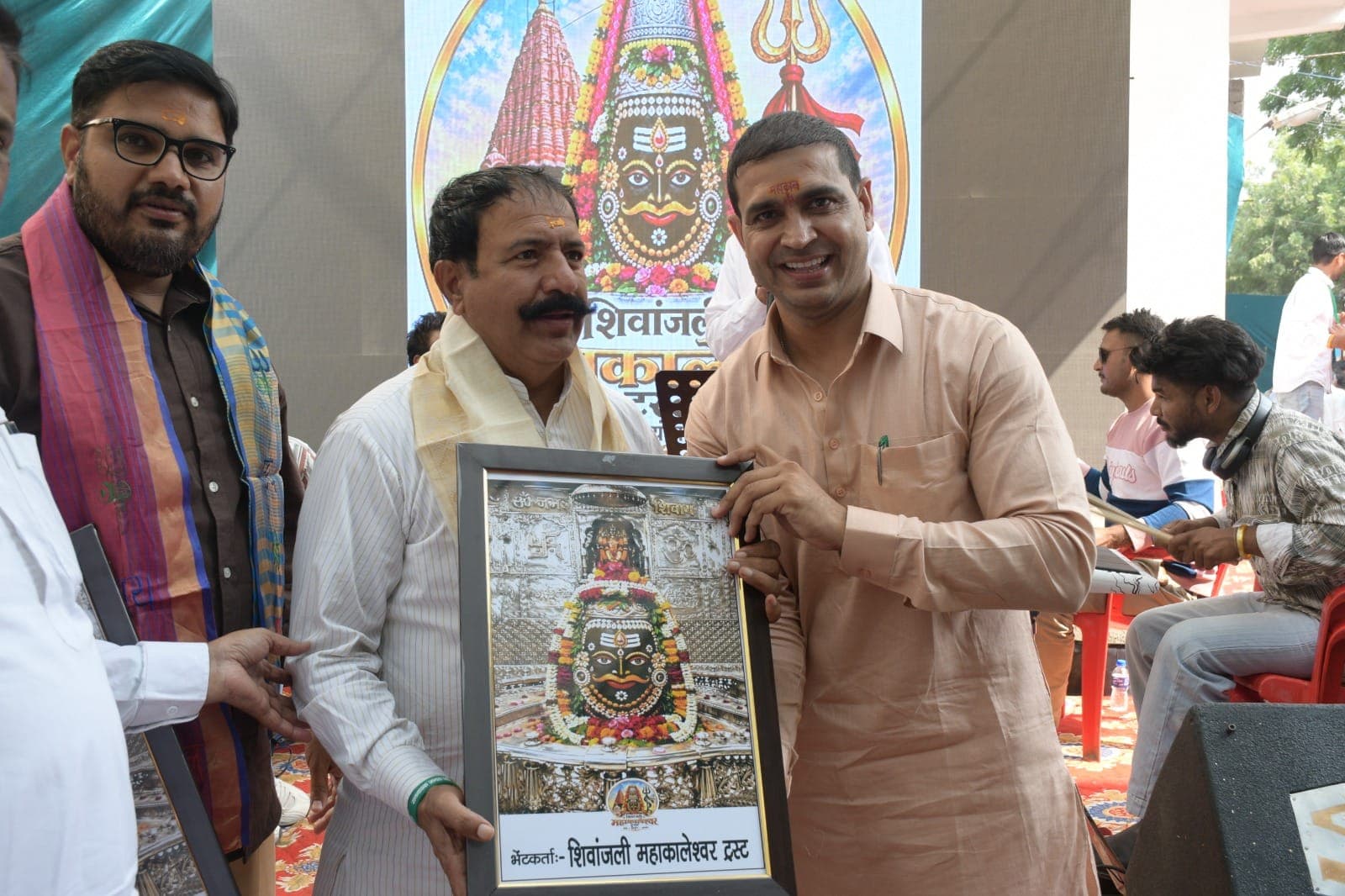 सफ़ेद धारीदार कुर्ते और क्रीम अंगवस्त्र में एक अतिथि को मंच पर देवता की फ्रेम वाली तस्वीर भेंट होती हुई
