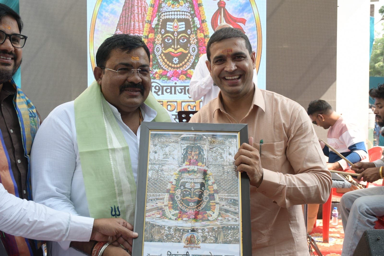 सफ़ेद कुर्ते और हरे अंगवस्त्र में एक अतिथि को मंच पर देवता की फ्रेम वाली तस्वीर भेंट होती हुई
