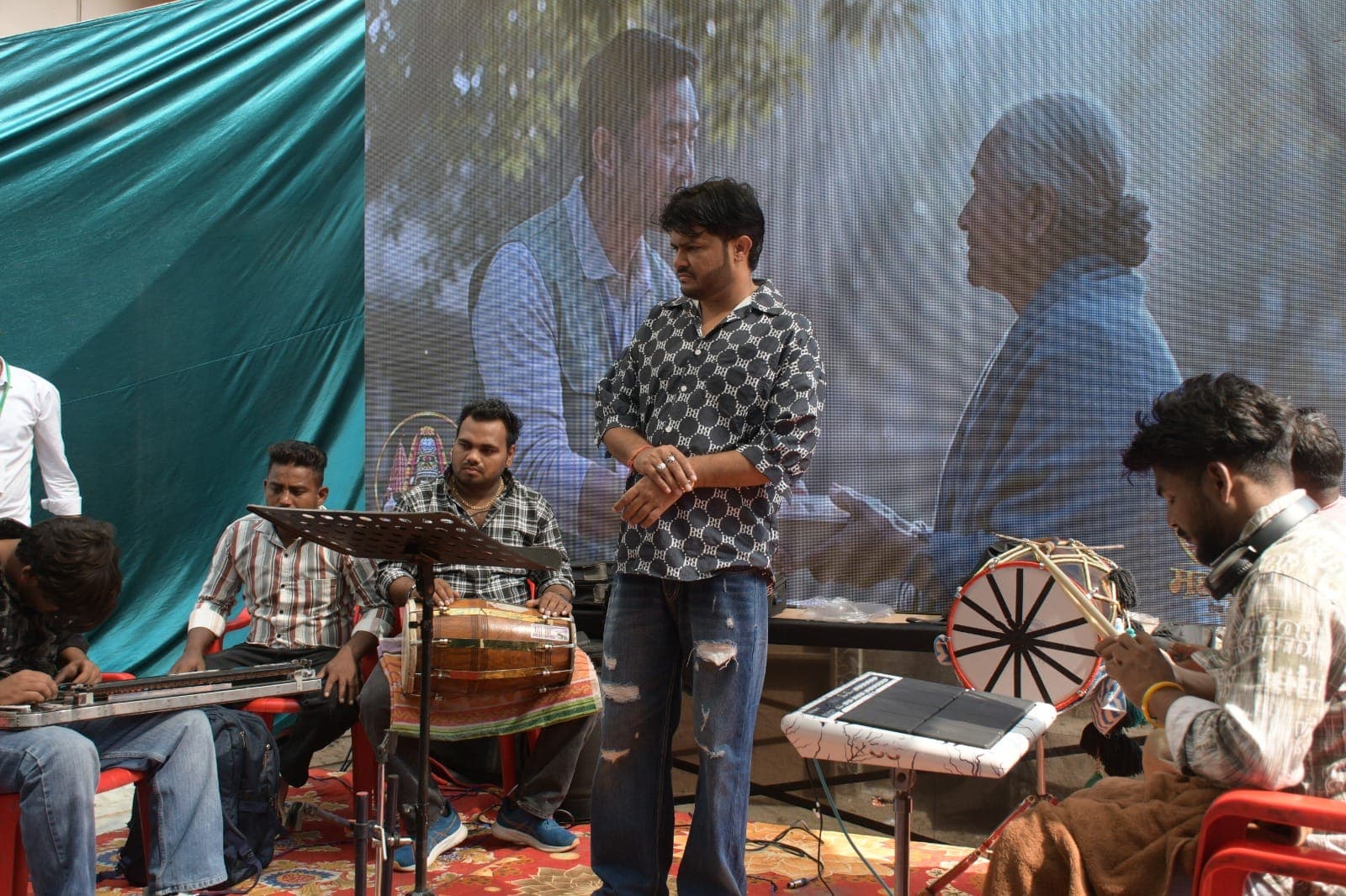 बड़ी वीडियो स्क्रीन के सामने मंच पर संगीतकार ढोलक, ढोल और अन्य वाद्य बजा रहे हैं