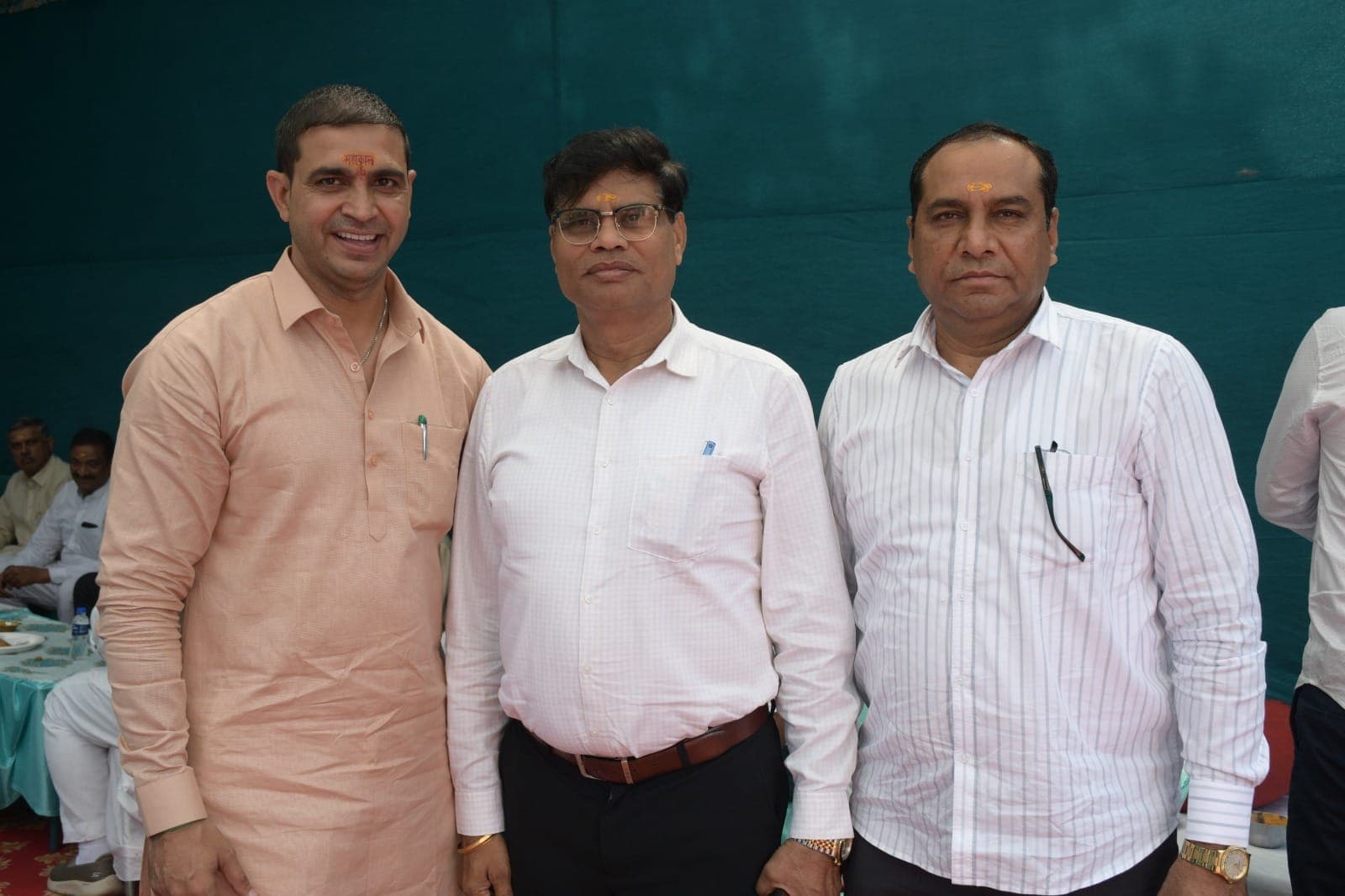 गहरे हरे-नीले पर्दे के सामने एक कुर्ता और दो सफेद कमीज़ पहने तीन पुरुष साथ खड़े हैं